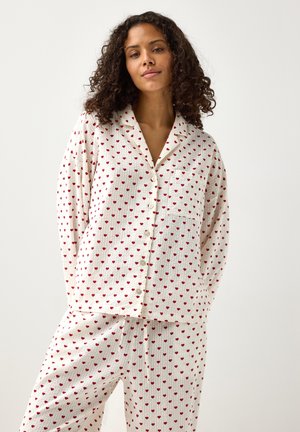 Vrouw met krullend haar die een witte pyjama met lange mouwen en knopen draagt, bedekt met kleine rode hartjes, staand tegen een effen achtergrond.