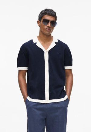 Granatowy krótki rękaw sweterkowej koszulki z kremowym kołnierzem i obszyciem. Posiada zapinany na guziki przód oraz małe logo wyszywane na piersi.