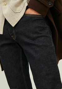 Os jeans de denim escuro têm costura amarela em contraste, um corte reto e uma textura subtil, combinados com um blaser leve em padrão espinha de peixe.
