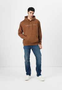 Bruine hoodie met witte logo, voorzak, ribgebreide manchetten en onderrand. Gecombineerd met blauwe spijkerbroek en witte sneakers op een neutrale achtergrond.
