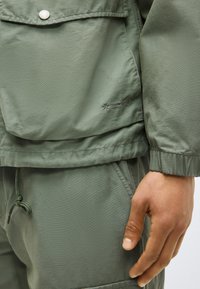 Veste verte avec poche à pression et poignet élastique, pantalon assorti avec taille à cordon, main reposant près de la poche.