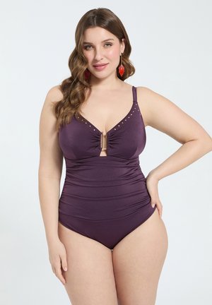 Kvinde med bølget brunt hår iført lilla heldragt med guldbelægning og central spænde, poserer med hånd på hoften mod hvid baggrund.