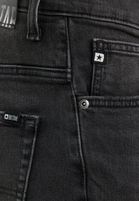 Fekete farmernadrág közeli képe, amely a hátsó zsebeket, övbújtatókat és csillaglogós márkajelzéseket, valamint egy fém szegecs részletet mutat.