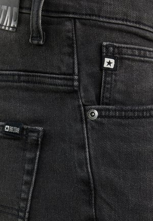 Close-up af sorte denimjeans, der viser baglommer, bæltestropper og mærkater med stjernelogos samt en metalnitte i detaljen.