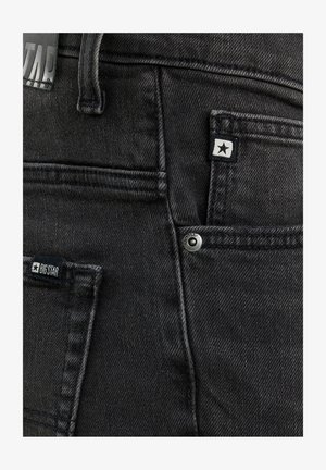 Krupni plan crnih traperica koji prikazuje stražnje džepove, petlje za remen i oznake marke sa zvjezdičastim logotipima te metalni detalj nitne.