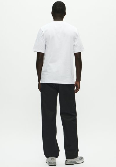 Mann som står med ryggen mot, iført en enkel hvit kortermet T-skjorte, svarte bukser og grå joggesko på en hvit bakgrunn.