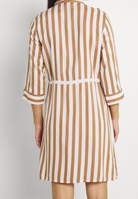 Pruhované šaty v hnědé a bílé barvě, s páskem v oblasti pasu, tříčtvrtečními rukávy a hladkým, jemným materiálem.