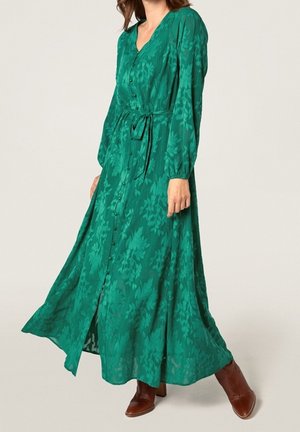 Kvinde iført en langærmet, grøn blomstret kjole med knapper og bånd i taljen, kombineret med brune ankelstøvler.