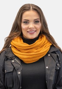Pletený hořčičný šál s texturovaným vzorem, omotaný několikrát kolem krku, spárovaný s černou bundou na zip.
