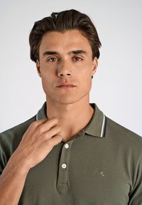 Olijfgroene polo met korte mouwen, twee knopen, contrasterende kraag met marineblauwe strepen, geborduurd logo op de linkerborst.