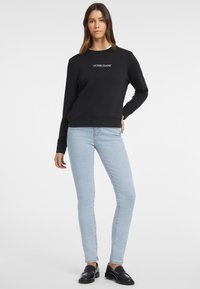 Felpa nera con logo "GUESS JEANS", collo a giro, e polsini a costine, abbinata a jeans skinny blu chiaro e mocassini neri.