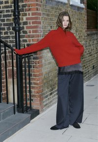 Rode gebreide trui met hoge hals, gecombineerd met een doorschijnend zwart topje en een wijde navy pantalon. Staande tegen een bakstenen muur, in een ontspannen pose.