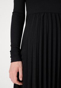 Mano descansando a un lado, vistiendo una camiseta de manga larga de canalé negra y una falda o vestido negro con pliegues, contra un fondo simple.