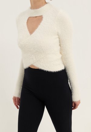 Kobieta ubrana w biały, puszysty, długi sweter z odsłoniętym dekoltem i czarne, wysokie, prążkowane spodnie z wysokim stanem.