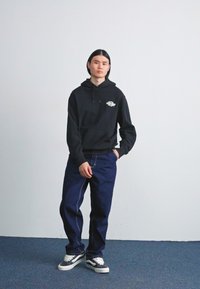 Sort hættebluse med en frontlomme og logo, parret med løstsiddende mørkeblå jeans og sorte og hvide sneakers på et blåt tæppe.