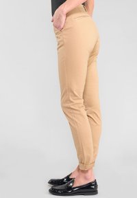 Seitenansicht einer Person, die beigen, tapered Hosen mit umgeschlagenen Säumen und schwarzen Lederloafers trägt, mit den Händen in den Taschen vor einem schlichten Hintergrund.