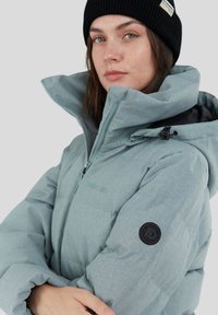 Chaqueta acolchada de color teal claro con cuello alto, cremallera y una insignia negra en la manga. Tejido texturizado con un acabado mate y capucha ajustable.