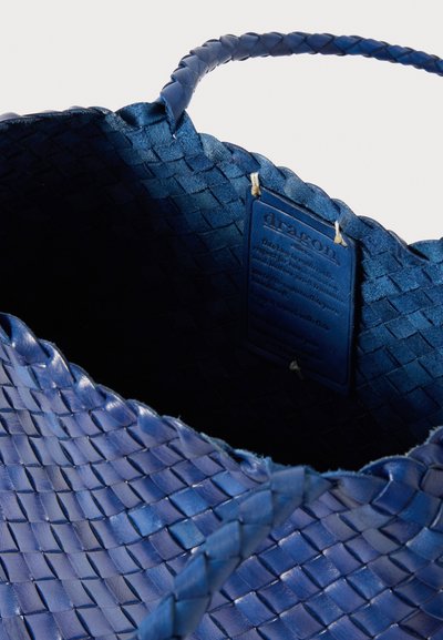 Πλεγμένη μπλε δερμάτινη τσάντα με υφή επιφάνειας, πλεγμένες λαβές και ετικέτα με οδηγίες φροντίδας μέσα.