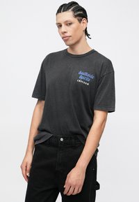 Melnas īsroku kokvilnas T-krekls ar zilā krāsā uzrakstu "Southside Spirits Chicago" kreisajā krūtīs, valkāts kopā ar melnām biksēm.