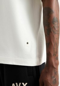 Camisa blanca de manga corta con un acabado texturizado, que presenta un pequeño detalle de ojal en el dobladillo, combinada con pantalones negros que muestran letras blancas.