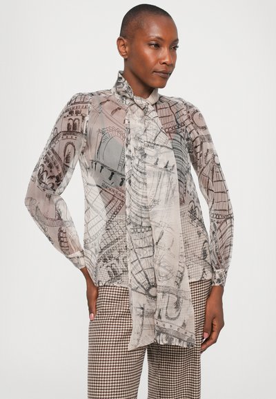 Femeie îmbrăcată cu o bluză transparentă cu mânecă lungă, decorată cu schițe arhitecturale negre și o cravată, asortată cu pantaloni cu model houndstooth maro și alb.
