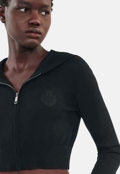 Felpa nera a maglia con design cropped, dotata di chiusura con zip e un sottile emblema ricamato sul petto. Tessuto morbido e stile aderente.