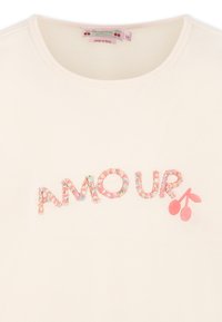 Beżowa koszula z napisem "AMOUR" wykonanym z wielokolorowych aplikacji w kształcie kwiatów, z małym akcentem w postaci różowej wiśni. Okrągły dekolt.