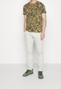 Tričko s kamuflážovým vzorem v zelené a hnědé barvě, doplněné o světle šedé slim-fit kalhoty a tmavě zelené semišové tenisky s šněrováním.