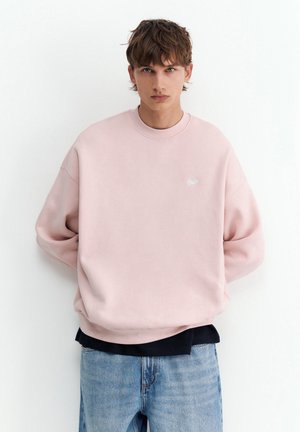 Jeune personne aux cheveux bruns portant un sweat-shirt ample rose clair et un jean bleu, debout contre un mur blanc.