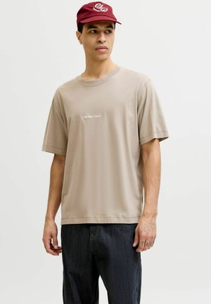 Uomo che indossa una camicia beige a maniche corte con piccoli testi bianchi e jeans scuri, guarda di lato, indossando un berretto rosso con logo bianco.