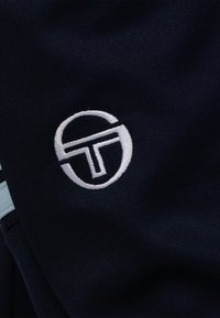 De marineblauwe stof heeft een wit geborduurd logo met een rond ontwerp en een "T"-detail. De textuur is glad met een lichte glans, versterkt door lichtblauwe accenten.