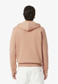 Sudadera con capucha de tela rosa suave con puños y dobladillo acanalados, que presenta un logo en la parte trasera, combinada con pantalones blancos. Textura suave y corte relajado.