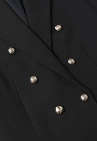 Tissu noir avec des boutons décoratifs dorés disposés en deux rangées verticales sur un vêtement, probablement un blazer ou un manteau.