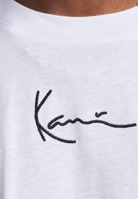 White cotton t-shirt featuring a black embroidered signature design. The fabric has a smooth texture and a crew neckline.