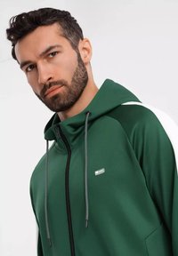 Felpa verde con zip e tessuto testurizzato, dotata di una cerniera nera e lacci, e dettagli bianchi sulle spalle. Presente un piccolo logo.