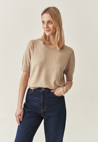 Beige kötött, rövid ujjú pulóver kerek nyakkal, sötétkék, magas derekú farmernadrággal párosítva. Simább textúra és finoman illeszkedő dizájn.
