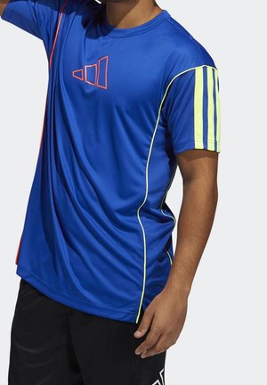 Mies, jolla on päällään sininen urheilupaita, jossa on keltaiset raidat hihoissa ja neonväriset reunukset, yhdistettynä mustiin shortseihin, yksiväristä taustaa vasten.