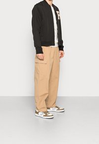 Svart bomberjacka med stickad krage och en tan-färgad "R" på bröstet, tillsammans med löst sittande beige cargo-byxor och bruna och vita sneakers.