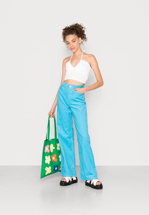 Jeans de perna larga azuis, top cropped branco com alças, sandálias plataformas pretas e uma mala de lona verde com padrões de flores coloridas.