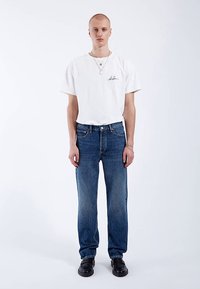 Valkoinen puuvillainen T-paita mustalla logolla, yhdistetty sinisiin farkkuihin. Asun täydentävät mustat kengät ja kerroksittain käytetyt hopeakorut.