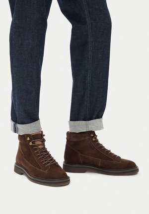 Person trägt dunkelblaue, umgeschlagene Jeans und braune Wildleder-Schnürstiefel und steht auf hellem Untergrund.