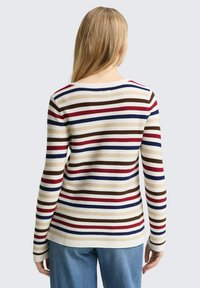 Vrouw met lang blond haar draagt een crèmekleurige trui met horizontale rode, marineblauwe, bruine en beige strepen, gecombineerd met lichtblauwe jeans.