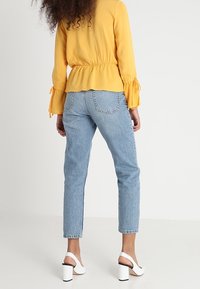 Blusa gialla con peplum, maniche lunghe e polsini a ruffle, abbinata a jeans a vita alta di colore blu chiaro e sandali bianchi con tacco a blocco.