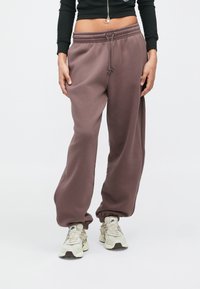 Asmuo vilkintis rudus laisvus sportinius kelnes, juodą trumpą užtrauktuką viršutine dalimi ir baltus Nike sportbačius, stovintis prieš paprastą fono.