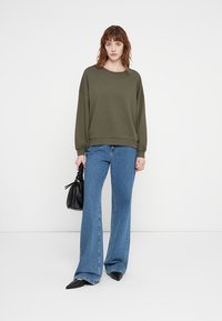 Khaki sweatshirt with wide sleeves and ribbed hem, paired with blue flared jeans and black pointed-toe flats. Holds a textured black bag.