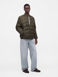 Мъж, облечен с olive green яке с подплата, широки светлосини дънки, тъмнокафяви обувки, стоящ с една ръка в джоба на обикновен фон.