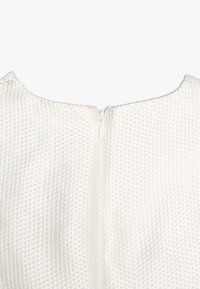 Witte textuurstof met een kruispatroon, voorzien van een centrale ritssluiting aan de achterkant. Gladde, afgeronde halslijn.