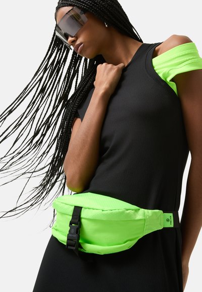 Neon groene heuptas van textuurstof, met een verstelbare zwarte gesp, gedragen over een zwart ribbeltop met contrasterende neonaccenten.