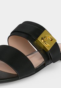 Μαύρο δερμάτινο σανδάλι με δύο λουράκια και μια χρυσή τετράγωνη αγκράφα με καμπύλη διακοσμητική λεπτομέρεια κλεισίματος.