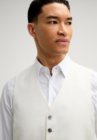 Weißer strukturierter Vest mit V-Ausschnitt, getragen über einem weißen Hemd mit Kragen. Verfügt über zwei dunkle Knöpfe und einen eleganten, minimalistischen Stil.